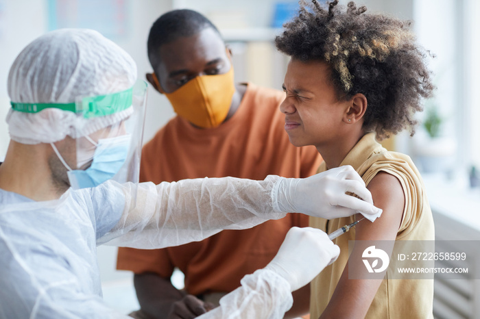 Portrait of teenage African-American boy squinting in pain during covid vaccination in clinic with father supporting him