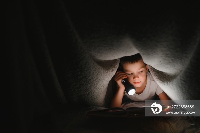 Boy with flashlight reading book under blanket at home