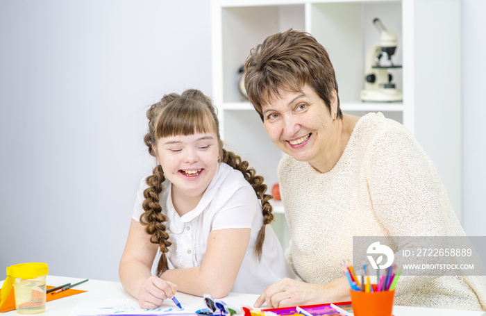 A girl with down syndrome is studying at home with her mother. Girl and mother sitting at the table draw a picture at home.