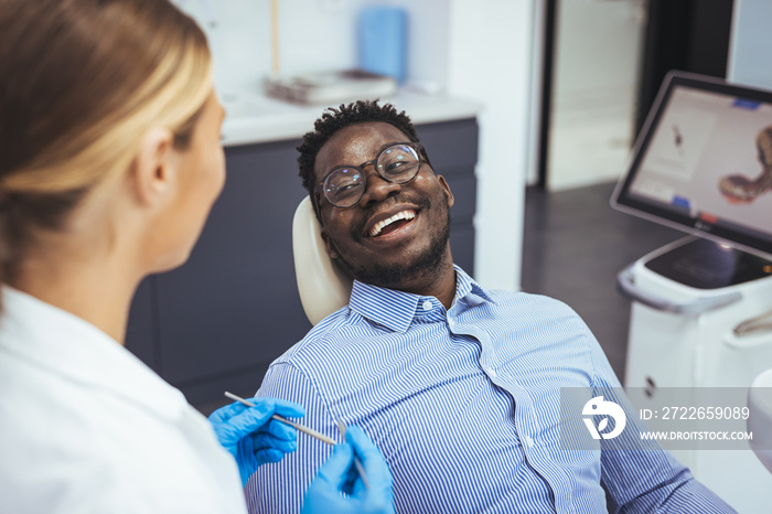 Female dentist with happy male patient at clinic. Happy patient sitting on chair while looking at dentist in medical clinic. Cheerful african american man with during examination in dental clinic