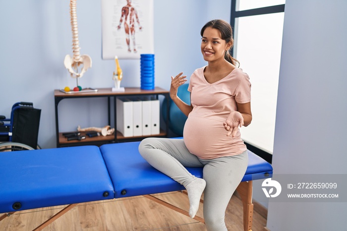 Young latin woman pregnant patient sitting on massage table at clinic