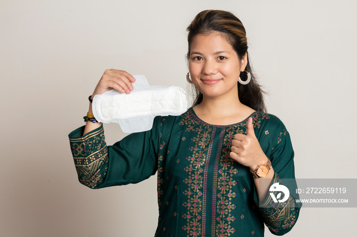 Young Indian girl holding a sanitary pad and showing thumbs up, wearing traditional dress, standing on a white isolated background.