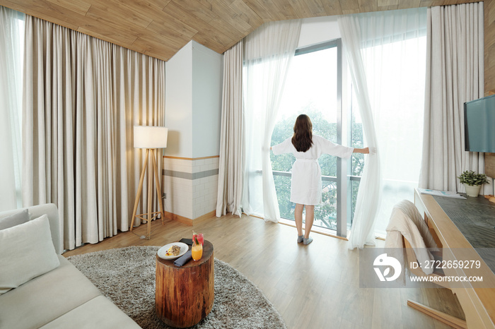 Young woman in robe opening curtains and looking outside in the morning, her breakfast is waiting her on coffee table, view from the back