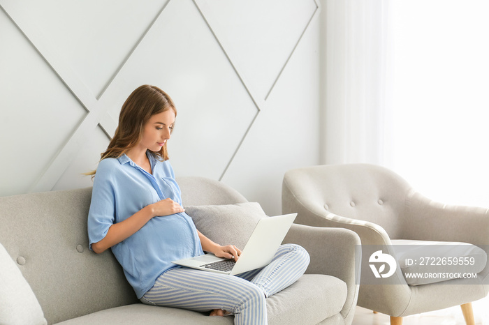 Beautiful pregnant woman with laptop sitting on sofa at home