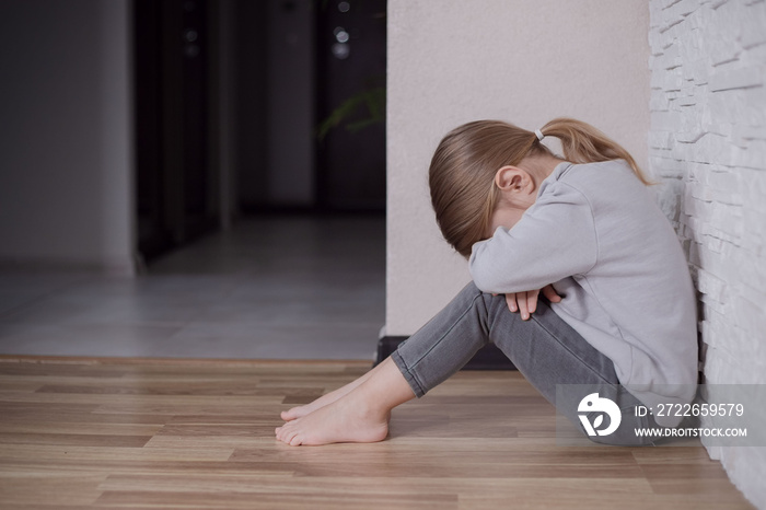 Sad little child girl sitting on floor in corner at home.