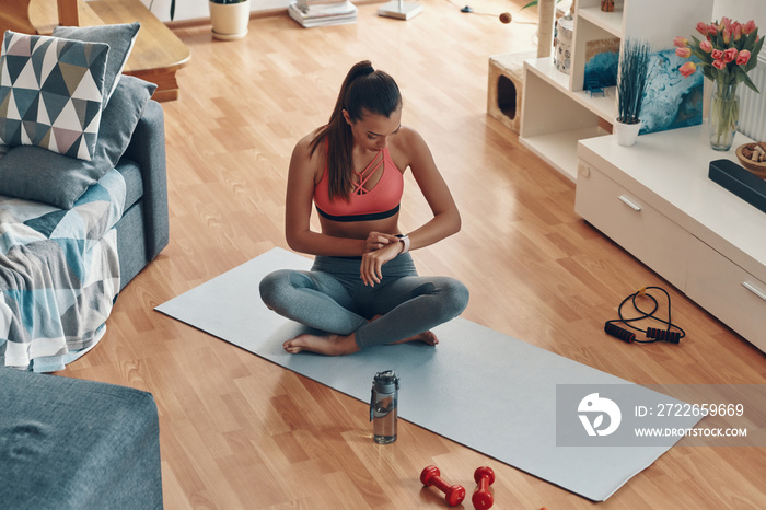 Top view of tired young woman in sports clothing checking her heart rate using smart watch while sitting in lotus position at home