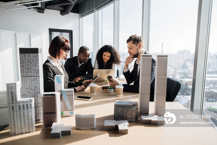 Business team of four good-looking multi ethnic corporate workers discussing financial report using laptop and gadgets working on building complex prototype project of residential or business district