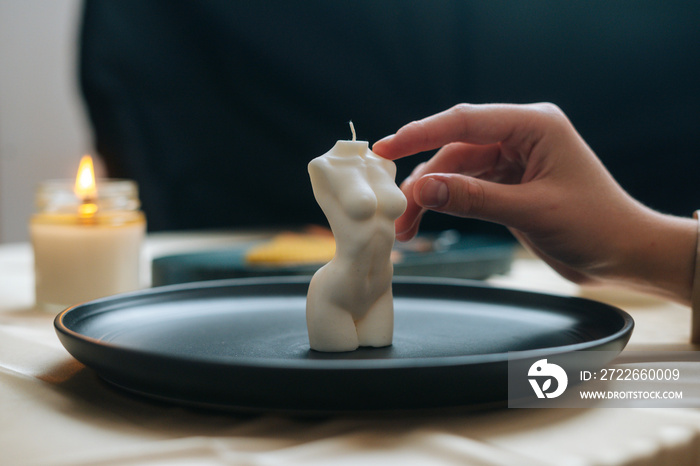 Close-up hands of unrecognizable female artisan putting on table ready to use handmade candle in shape of body of young woman, candles burning around. Process of making handmade natural candles.