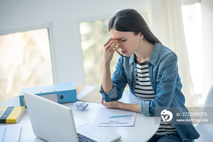 Upset dark-haired female sitting at her desk with unemployment claim, touching her nose bridge