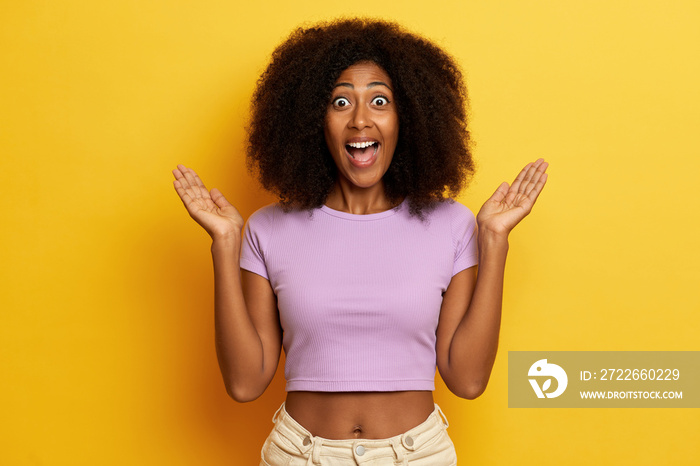 Surprised curly haired woman smiling toothily with opened wide mouth, spreads palms aside, posing over yellow background.