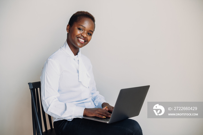 Cheerful African-american woman using laptop, having online meeting, sitting on chair against white wall. Looking and smiling at camera. Laughing confident