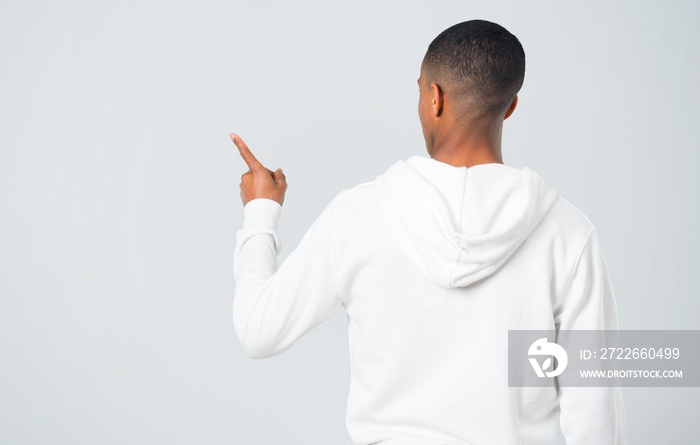 Dark-skinned young man with white sweatshirt pointing back with the index finger on grey background