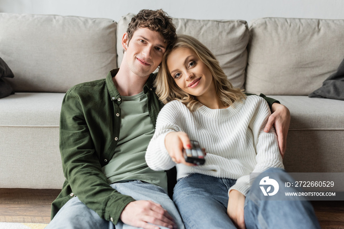 happy blonde woman clicking channels with remote controller near boyfriend in living room.