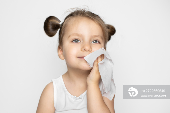 Little beautiful Caucasian blond girl with ponytails, wipes her face and cheek with a wet napkin, looking up and smiling. Studio shot on isolated white background with copy space