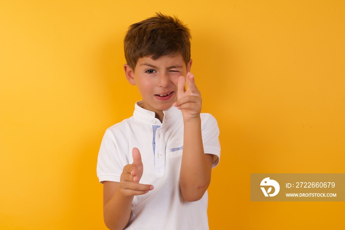 Hey you, bang. Joyful and charismatic good-looking little cute boy kid wearing white t-shirt against yellow wall winking and pointing with finger pistols at camera happily and cheeky.