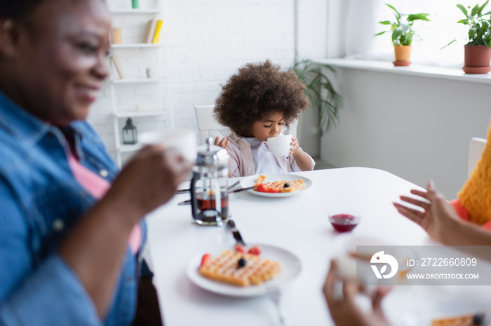 african american child drinking tea during breakfast with blurred granny and mom