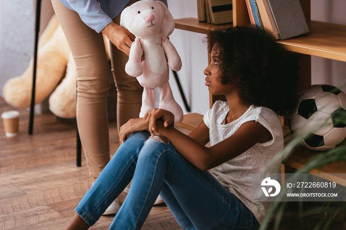 Upset african american girl with autism looking at soft toy in hands of psychologist standing near on blurred foreground