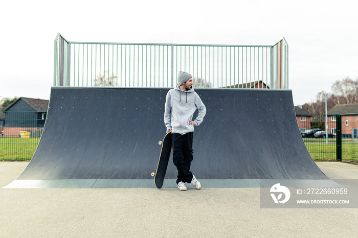 Portrait of a young adult male with a skateboard standing in front of a large ramp at a local skatepark. Skateboarding healthy lifestyle concept