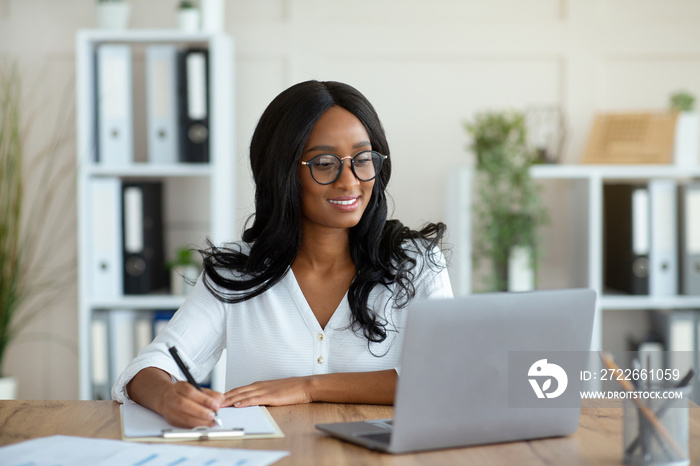 Cheerful black business lady working on laptop, taking notes during online work meeting or webinar at office