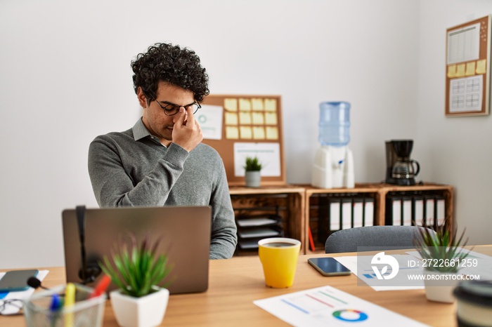 Young hispanic man wearing business style sitting on desk at office tired rubbing nose and eyes feeling fatigue and headache. stress and frustration concept.