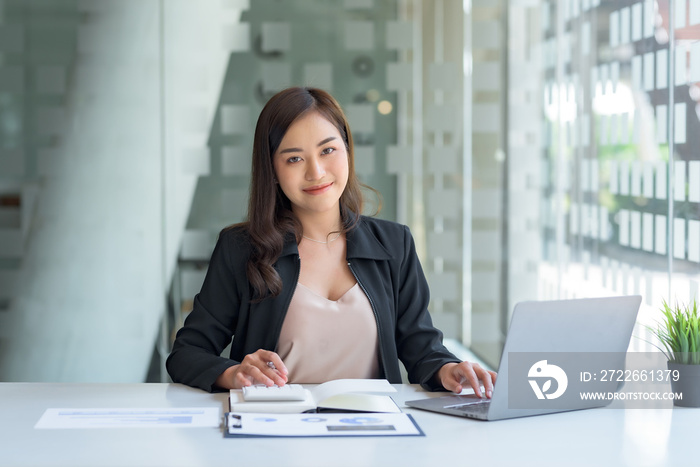 Smiling beautiful Asian businesswoman sitting at work looking at camera using laptop and computer while taking notes and working on documents at the office.