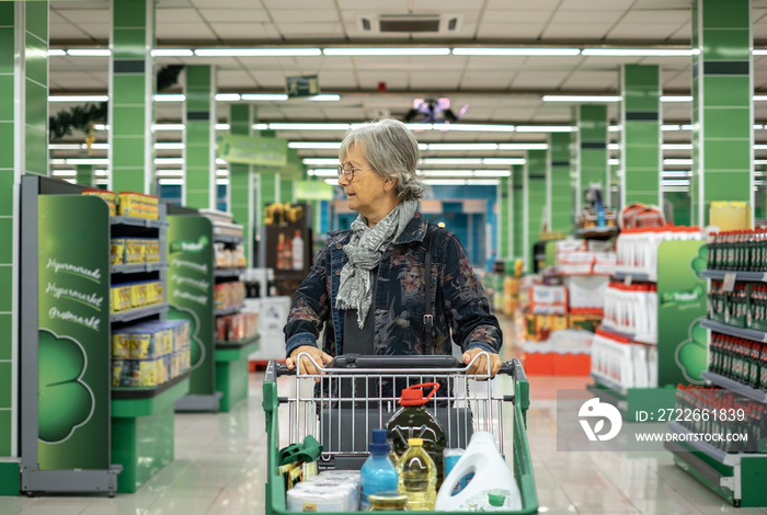 Caucasian senior woman pushing cart in supermarket looking at the products on display, consumer purchase concept