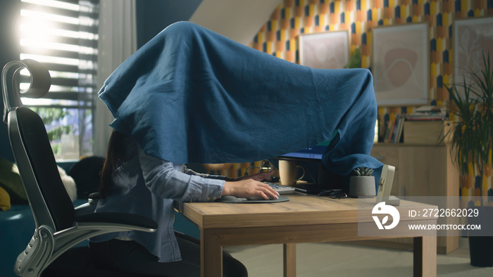 Woman sitting on chair at the table at home and doing color correction and grading, hiding under the blanket from sunlight rays of sun which comes from window