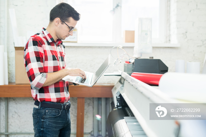 Side view of casual smart man in glasses working with laptop in modern office with printing equipment