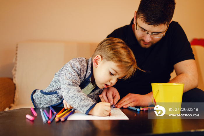 Small boy and his uncle or father sitting by the table at home playing with crayons color pencils drawing and learning family activities having fun