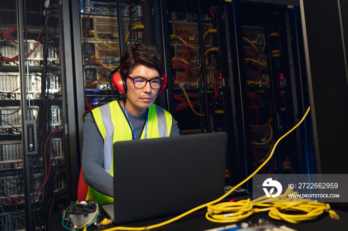 Asian male engineer wearing ear plugs using a laptop in computer server room