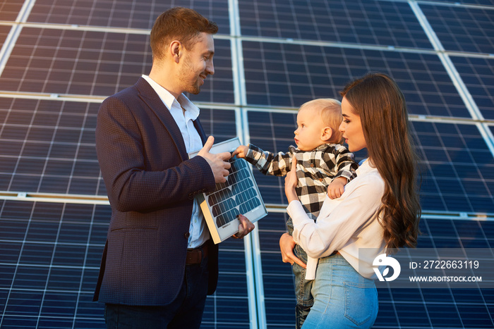 Cheerful kid in his mother arms examines the solar battery that dad holds, against background solar panels. Concept of family warmth and comfort in modern world with continuously advancing technology