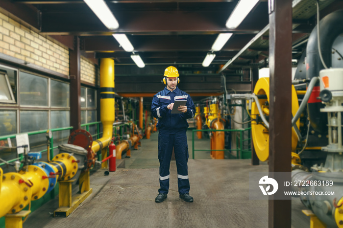 Full length of caucasian worker in protective suit, helmet and antifones on ears using tablet for work while standing in heavy industry plant.