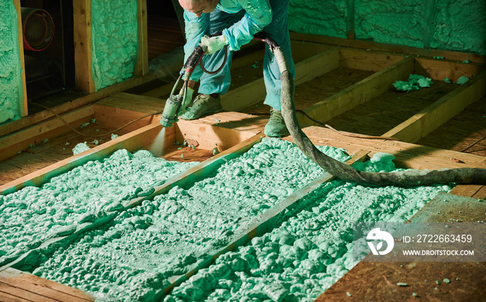 Male builder insulating wooden frame house. Cropped view of man worker spraying polyurethane foam on floor inside of future cottage, using plural component gun. Construction and insulation concept.