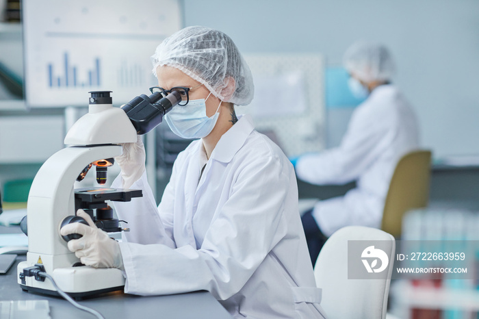 Young doctor in mask and lab coat looking through the microscope at the table, she examining medical samples