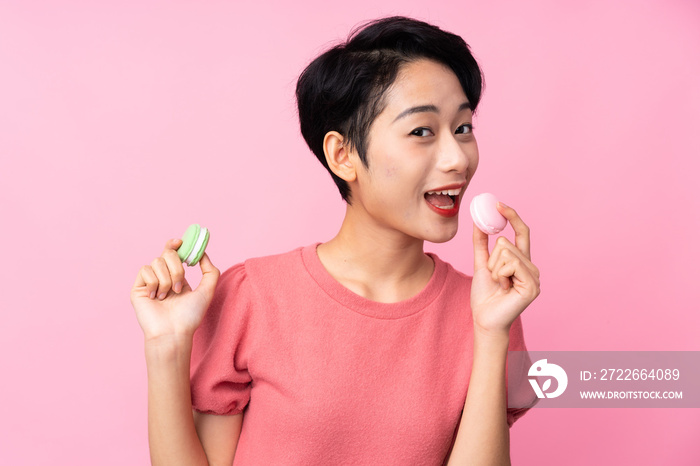 Young Asian girl over isolated pink background holding colorful French macarons and eating it