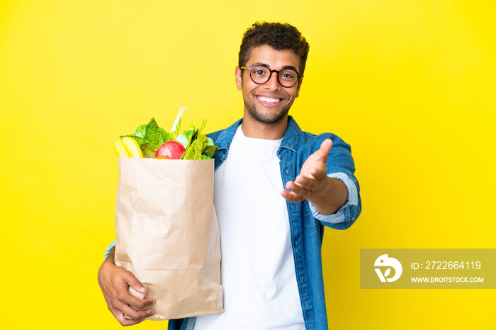Young Brazilian man holding a grocery shopping bag isolated on yellow background shaking hands for closing a good deal