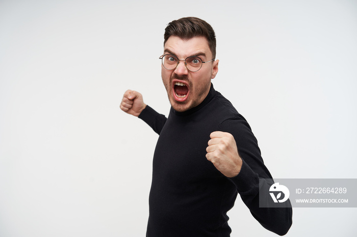 Angry young handsome brunette male with short haircut screaming madly and clenching raised hands in fists while going to hit someone, isolated over white background