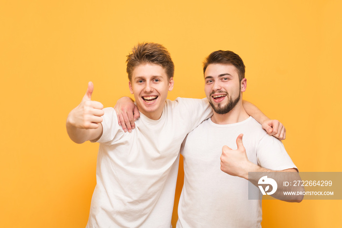 Portrait of a two happy young men showing thumbs up isolated over yellow background. Two happy friends in white t-shirts hugged by men, looking at the camera, smiling and showing a thumbs up