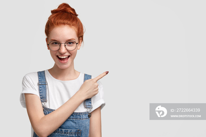 Joyful redhead female model with hair bun, glad to demonstrate her project work, indicates with fore finger aside, dressed in fashionable outfit, has positive expression, isolated over white wall