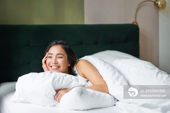 Happy mornings. Beautiful korean woman lying in bed, smiling, waking up upbeat, cover herself with warm duvet