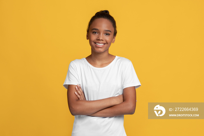 Happy calm teen black girl pupil in white t-shirt with crossed arms, isolated on yellow background