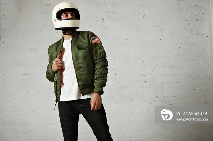 Proud looking motorbiker in a plain white helmet, khaki bomber jacket, jeans and t-shirt against white wall background