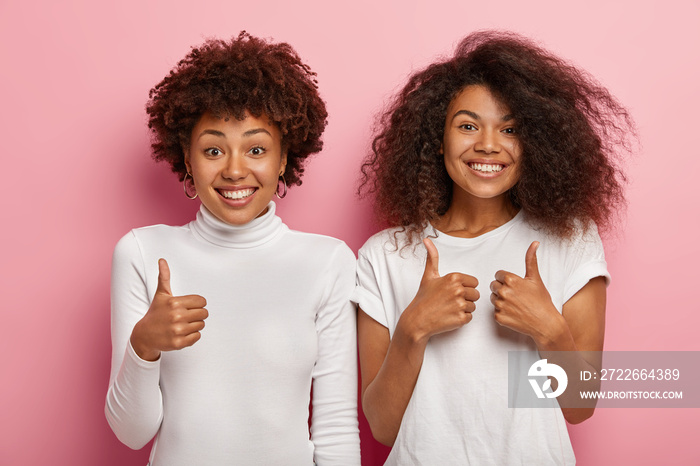 Glad women best friends make thumb up sign, smile cheerfully, show support and agreement, like something, wear white casual t shirt and turtleneck, isolated on pink background, say its cool.