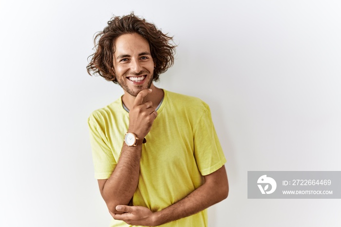 Young hispanic man standing over isolated background looking confident at the camera smiling with crossed arms and hand raised on chin. thinking positive.