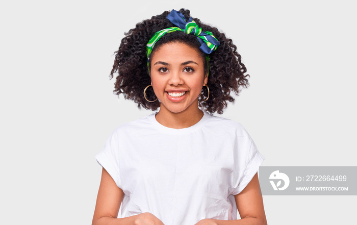 Beautiful happy dark-skinned young woman dressed in white t-shirt, feeling happy. African American female smiling broadly, wearing white t-shirt and trandy headband posing over white studio wall