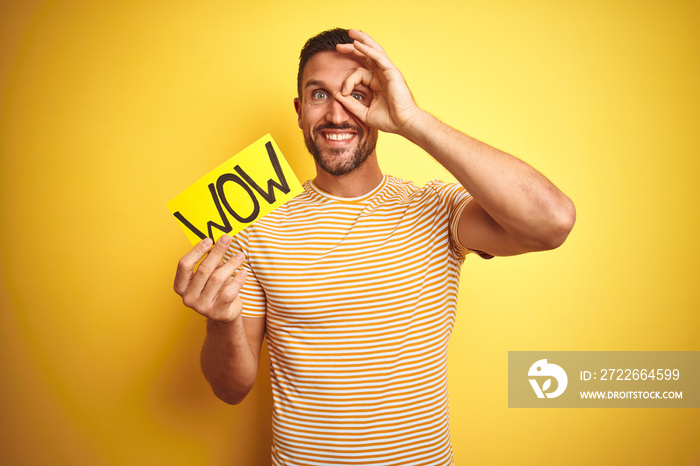 Young man holding wow text banner over yellow isolated background with happy face smiling doing ok sign with hand on eye looking through fingers