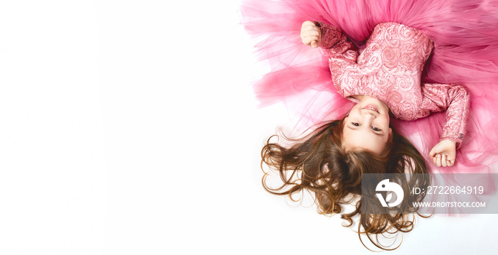 little Princess. Portrait of a cute Caucasian little laughing girl in an evening bright pink dress lies on a white background. Horizontally. top view