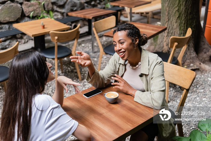 African american woman talking to friend near coffee and smartphone on terrace of cafe