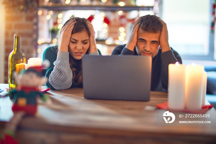 Young beautiful couple sitting using laptop around christmas decoration at home suffering from headache desperate and stressed because pain and migraine. Hands on head.