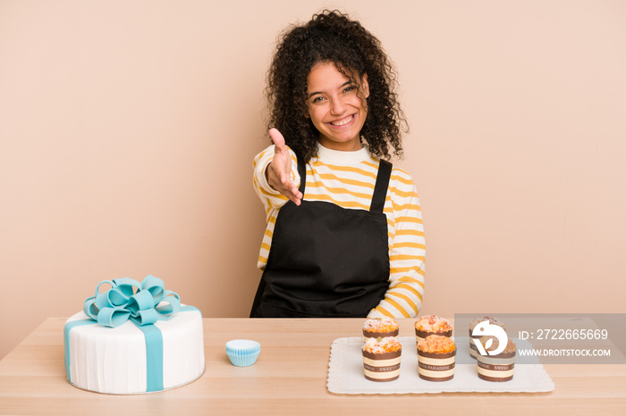 Young african american woman preparing a sweet cake and muffins on a table stretching hand at camera in greeting gesture.